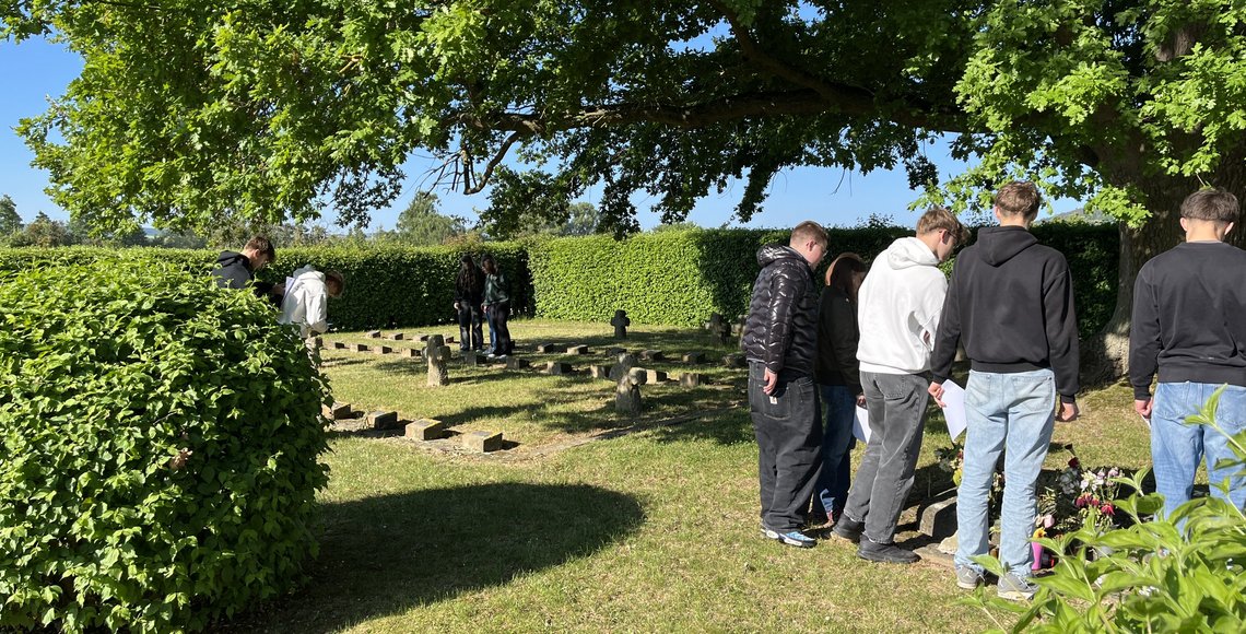 Auf dem Bild sind Schülerinnen und Schüler auf der KRiegsgräberstätte zu sehen. Sie stehen in kleinen Gruppen zusammen und lesen die Inschriften auf den Gräbern und auf einem Gedenkstein.